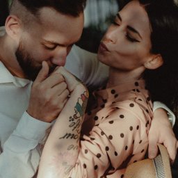 Red Dot Kamil Matuszewski - Zakochana para przytulona w plenerze: mężczyzna w białej koszuli całuje dłoń kobiety w różowej sukience w grochy, widoczny tatuaż na ramieniu kobiety i słomkowy kapelusz.