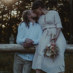 Red Dot Kamil Matuszewski - Para całująca się na tle zalesionego krajobrazu, ona w zwiewnej sukience i botkach trzyma bukiet suszonych kwiatów, on w białej koszuli i spodniach khaki, opierają się o drewniany płot.