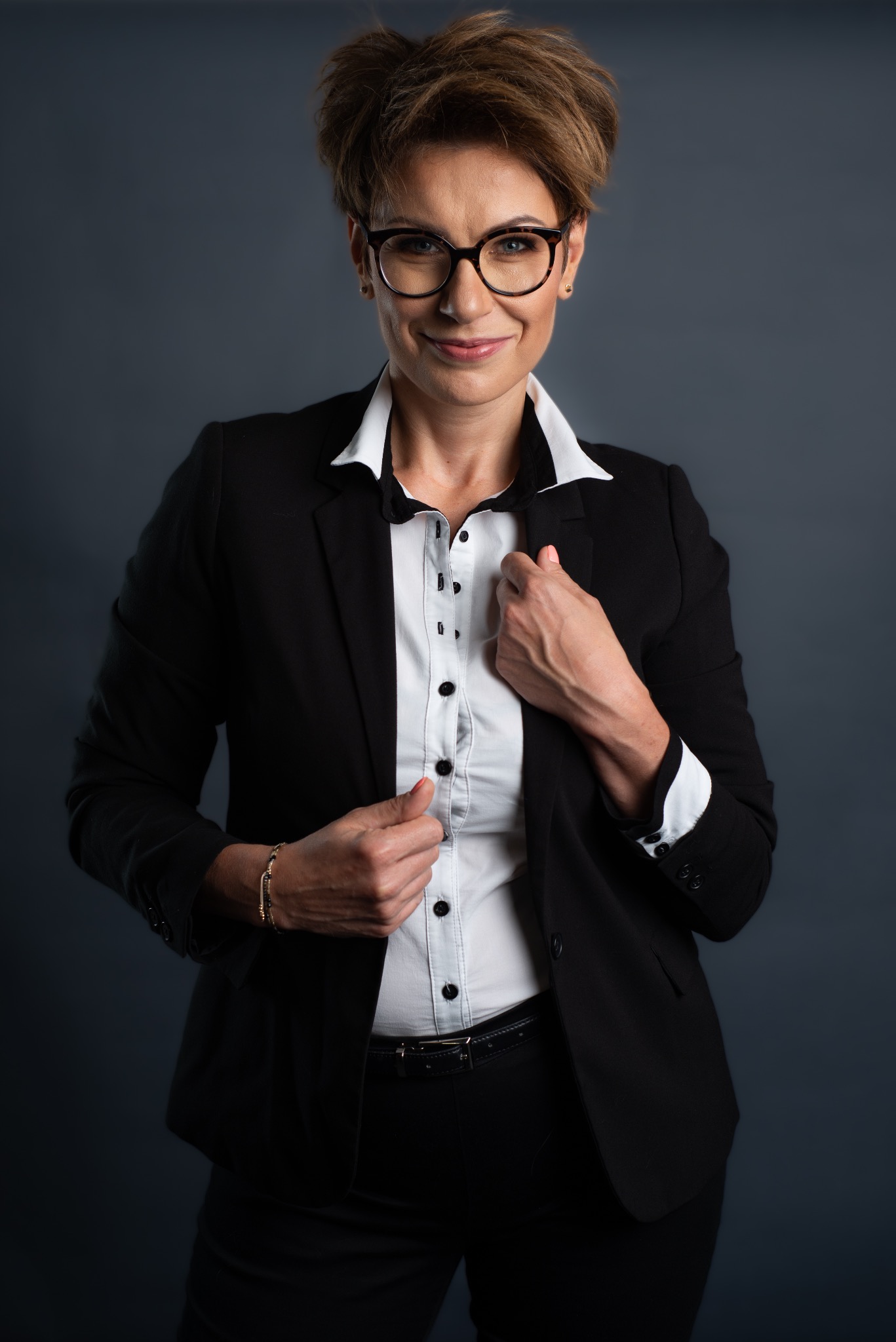 Portret kobiety w okularach, ubrana w czarny garnitur i białą koszulę, pozująca na szarym tle.