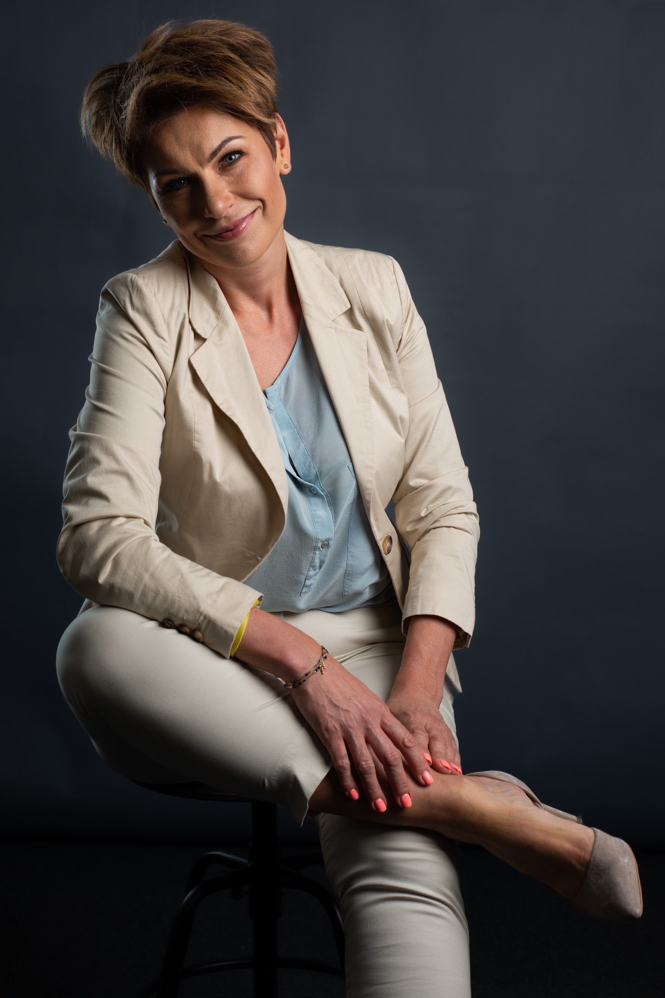 Portret kobiety w beżowym garniturze, siedzącej na krześle na ciemnym tle, z uśmiechem i skrzyżowanymi nogami.
