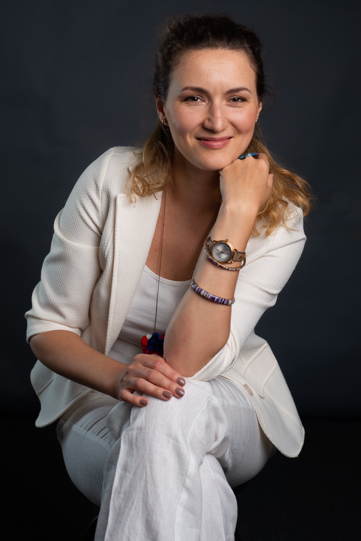 Portret uśmiechniętej kobiety w białym stroju, z zegarkiem i bransoletką z fioletowymi akcentami, opierającej podbródek na dłoni na ciemnym tle.