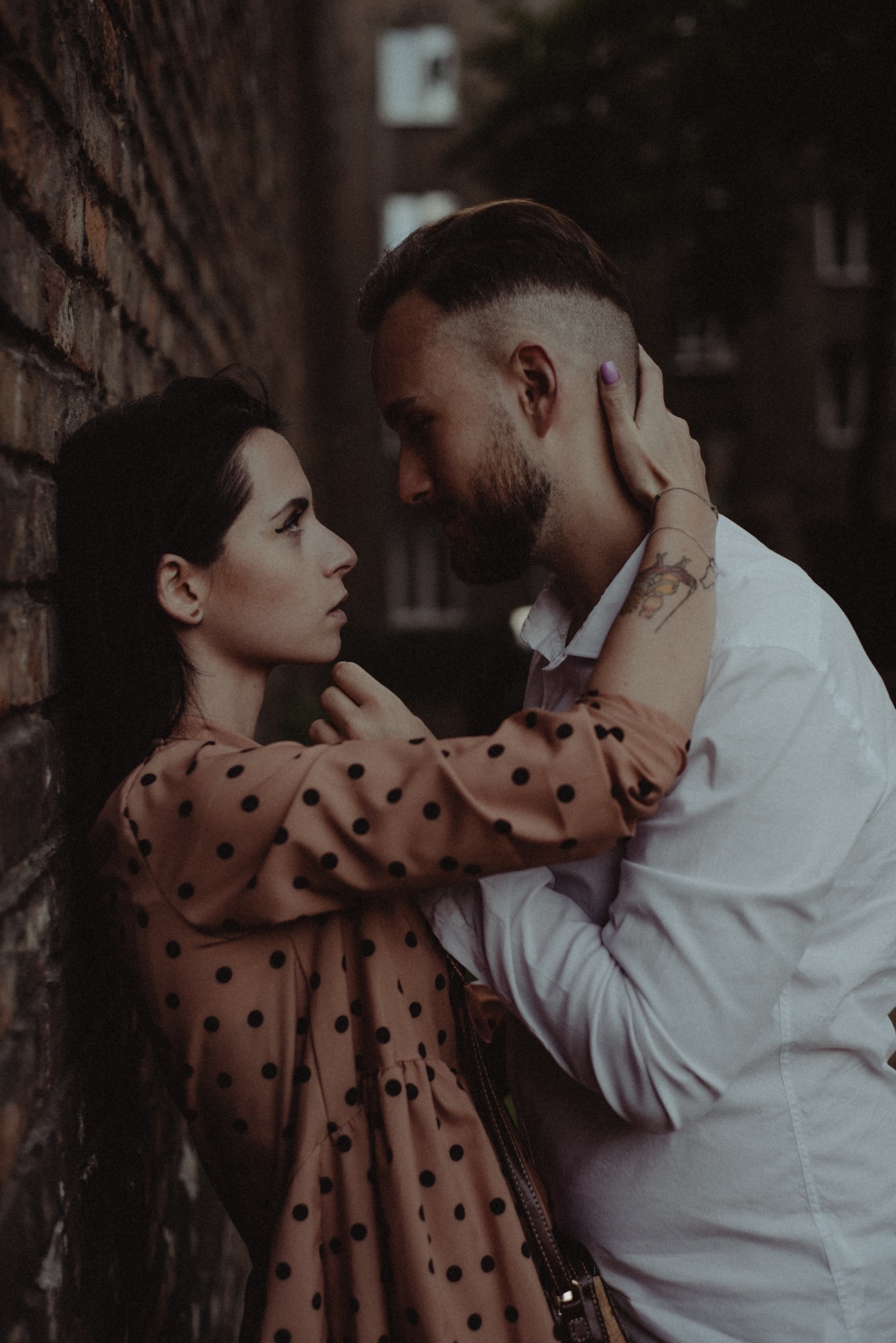 Para zakochanych, kobieta w sukience w grochy oparta o ceglaną ścianę, mężczyzna w białej koszuli obejmuje ją, wzajemne spojrzenia, tatuaż na ramieniu mężczyzny.