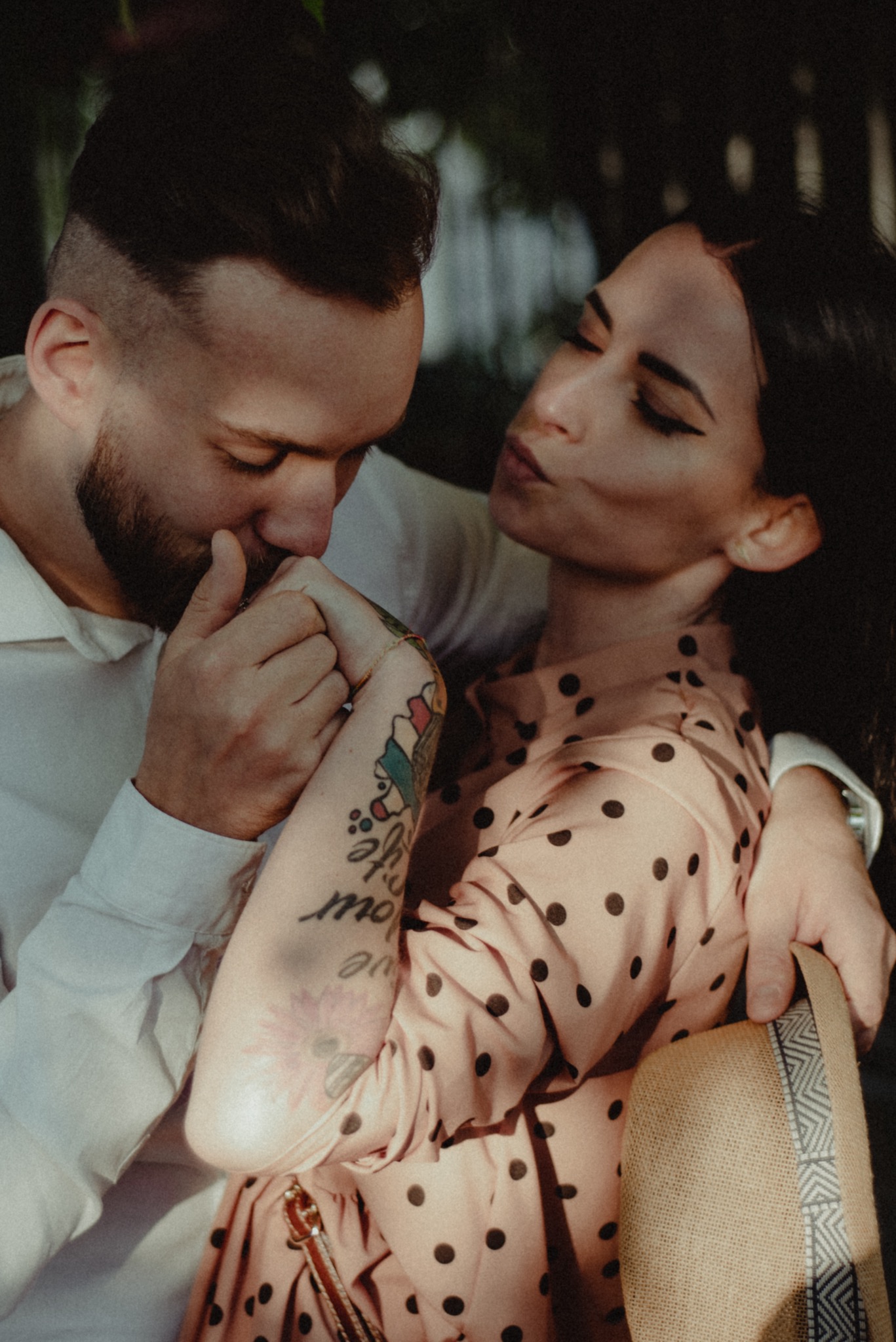 Zakochana para przytulona w plenerze: mężczyzna w białej koszuli całuje dłoń kobiety w różowej sukience w grochy, widoczny tatuaż na ramieniu kobiety i słomkowy kapelusz.