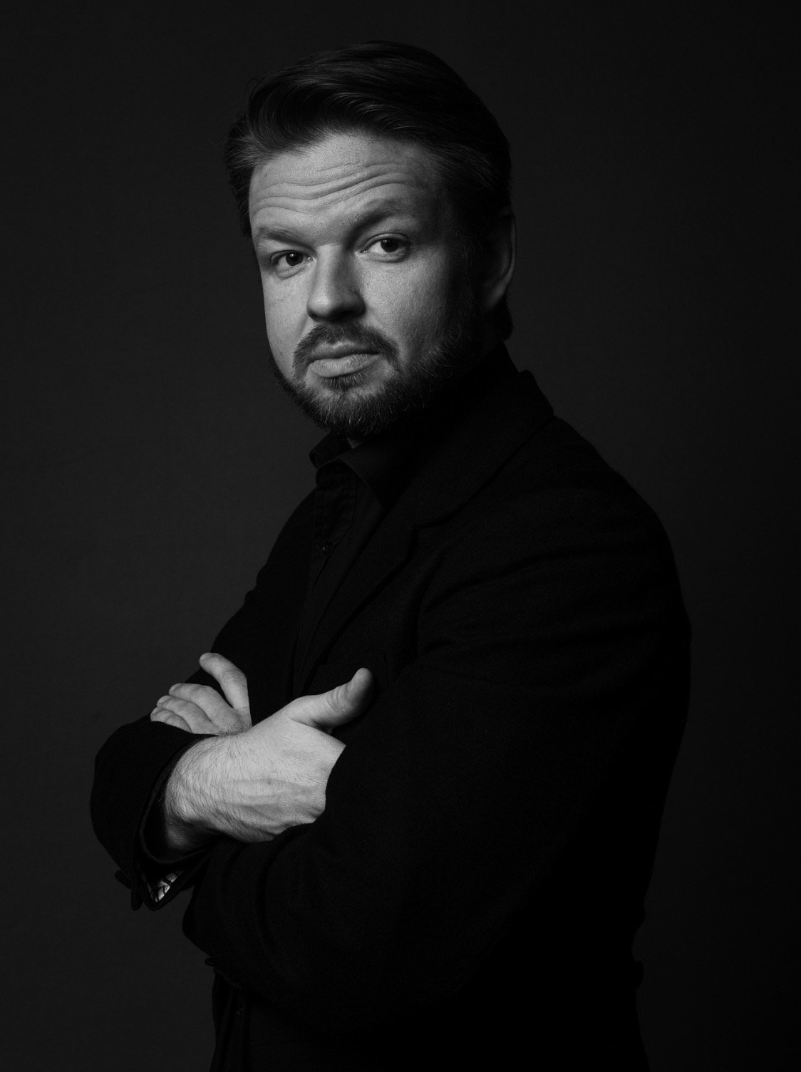 Portret biznesowy mężczyzny w czerni, z brodą, na ciemnym tle, z założonymi rękami.