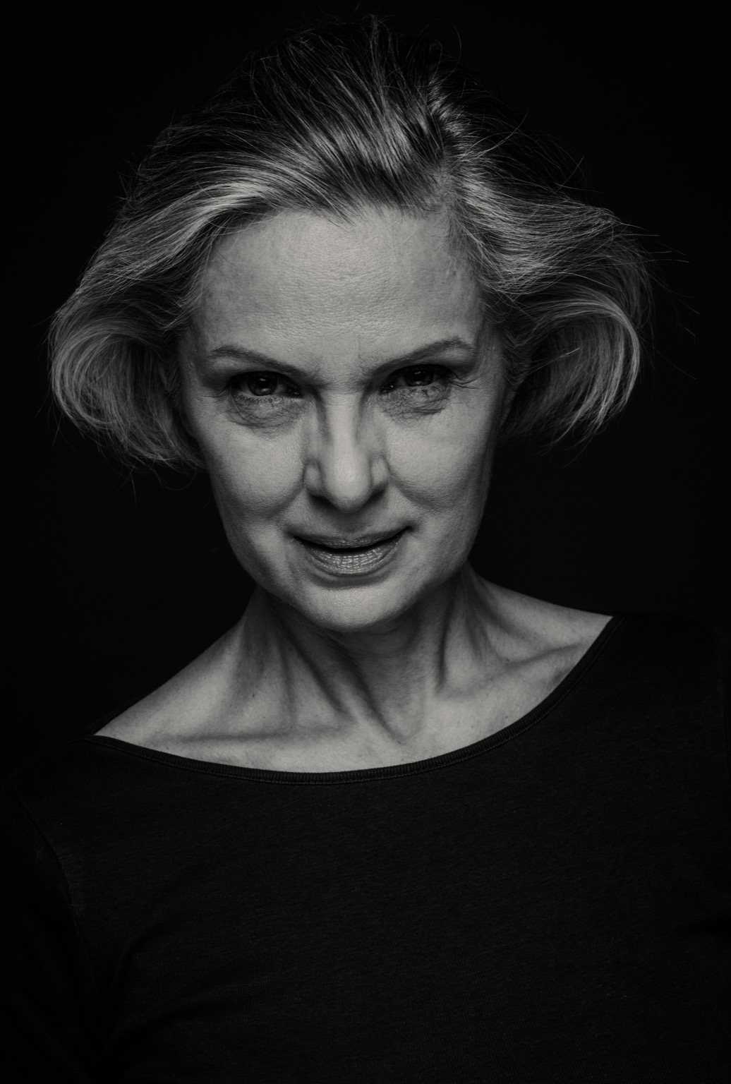 Portret kobiety w średnim wieku, czarno-biały, z krótkimi, siwiejącymi włosami, ubrana w czarną bluzkę z szerokim dekoltem, na ciemnym tle.