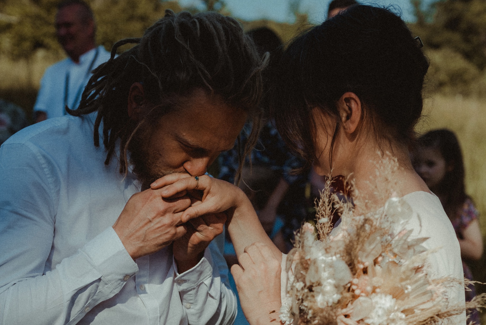 Pan z dredami w białej koszuli całuje dłoń panny młodej z bukietem polnych kwiatów, w tle zamazane postacie gości.