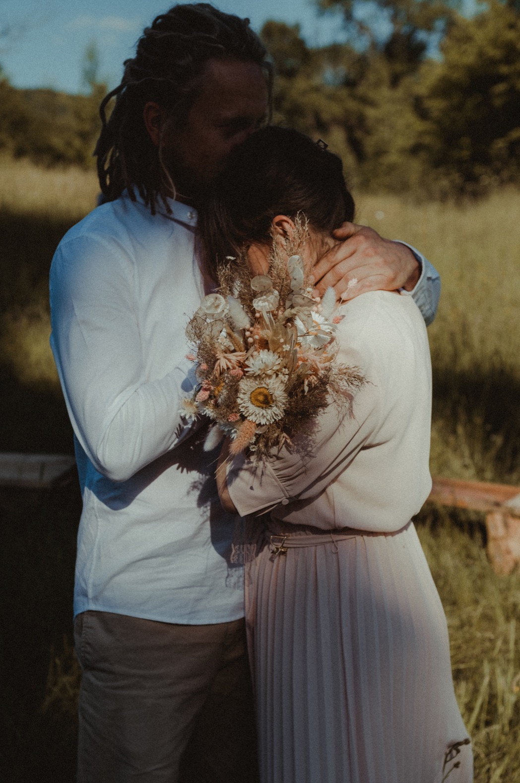 Mężczyzna z dredami obejmuje kobietę trzymającą bukiet polnych kwiatów, delikatne światło pada na parę na tle łąki.