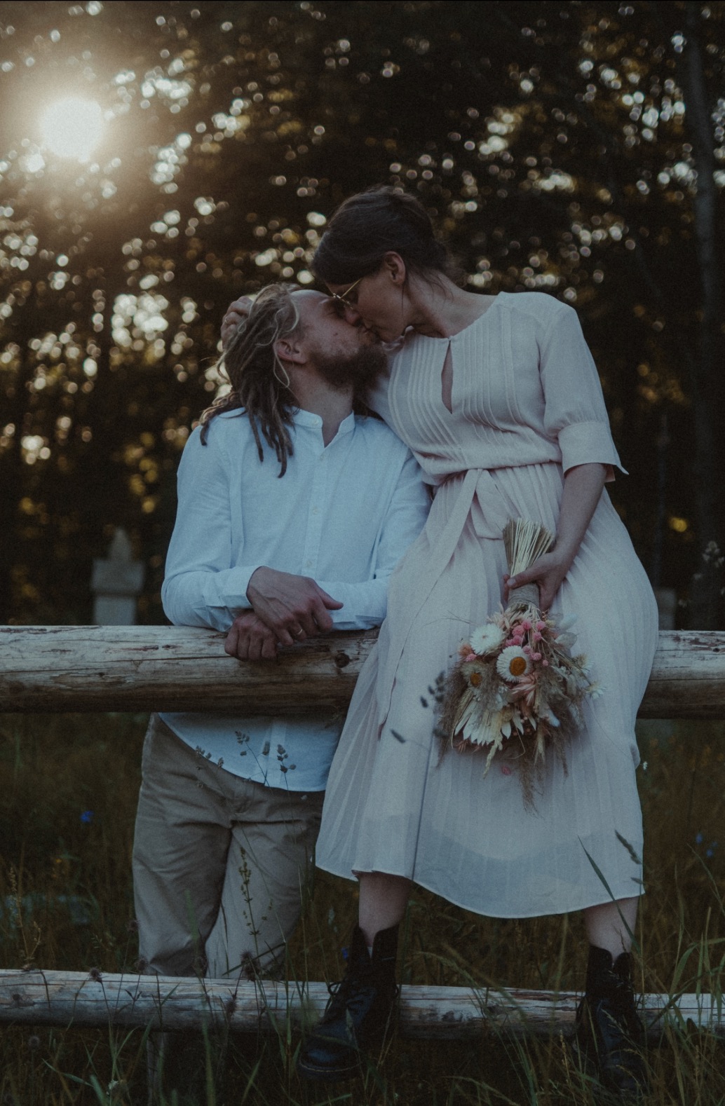 Para całująca się na tle zalesionego krajobrazu, ona w zwiewnej sukience i botkach trzyma bukiet suszonych kwiatów, on w białej koszuli i spodniach khaki, opierają się o drewniany płot.
