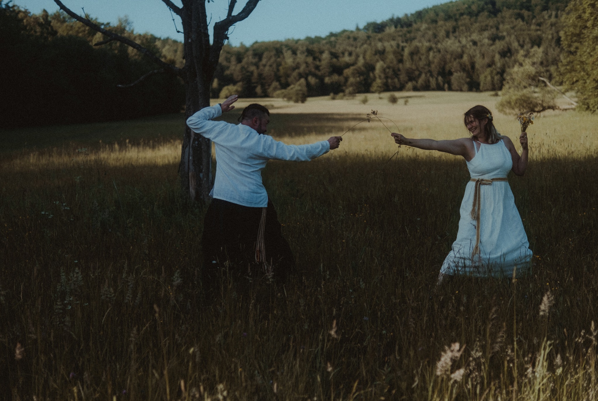 Para w białych strojach bawi się na łące, trzymając gałązki, z drzewem i lasem w tle.
