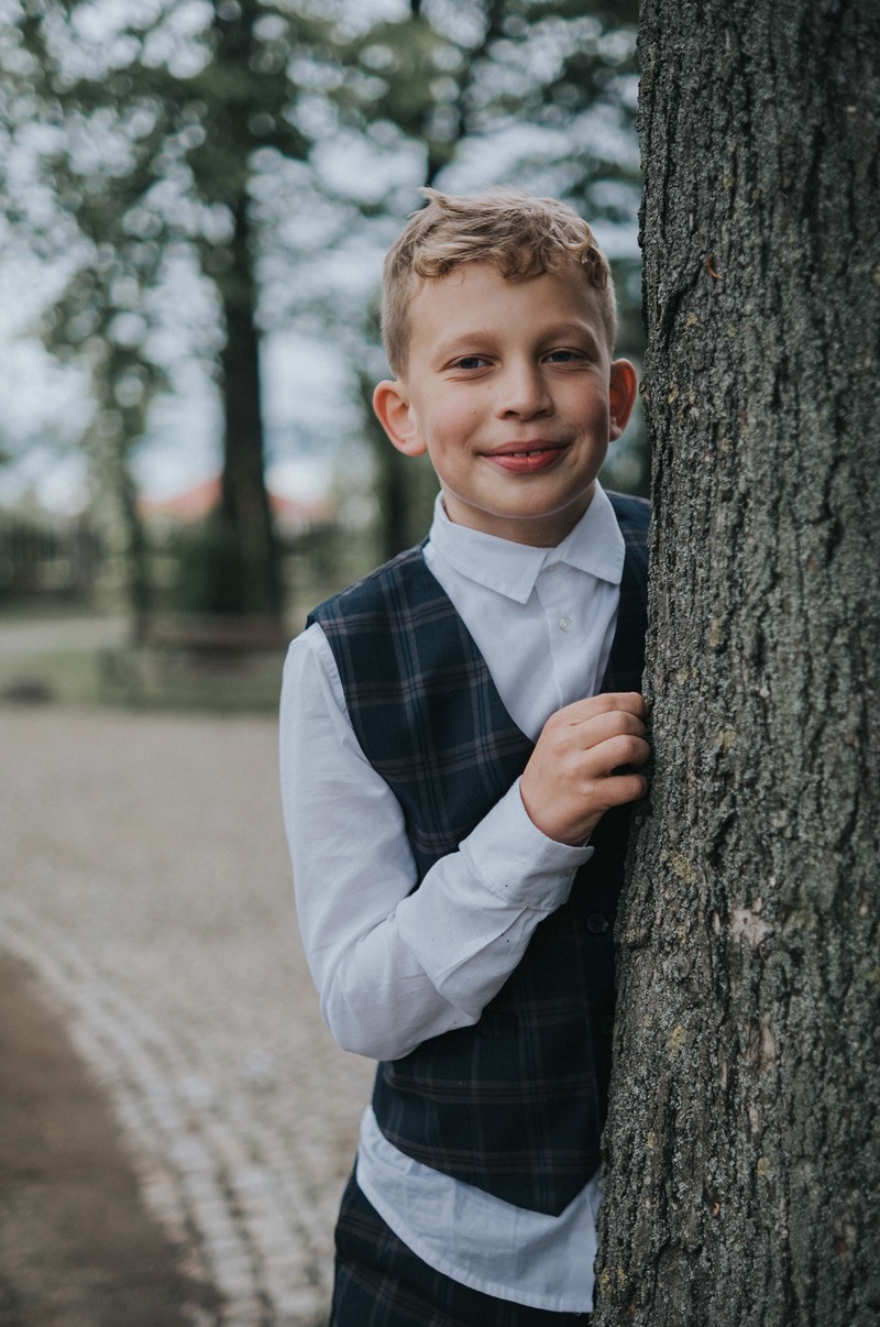 Uśmiechnięty chłopiec w eleganckiej kraciastej kamizelce i białej koszuli, oparty o korę drzewa na zewnątrz.