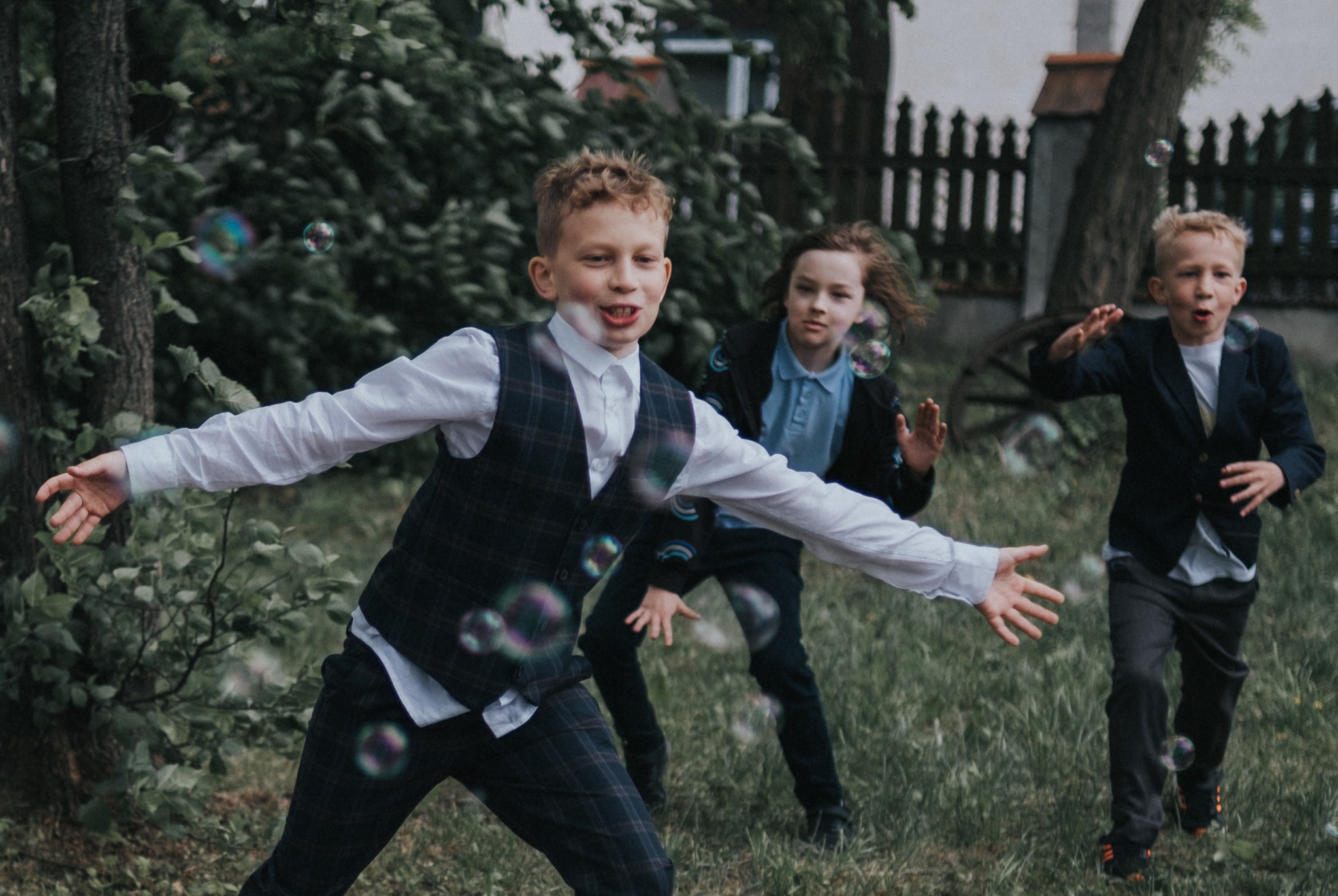 Trzech chłopców w eleganckich strojach bawi się na trawie puszczając bańki mydlane, jeden z nich wyciąga ręce, próbując je złapać, w tle drzewa i zieleń.