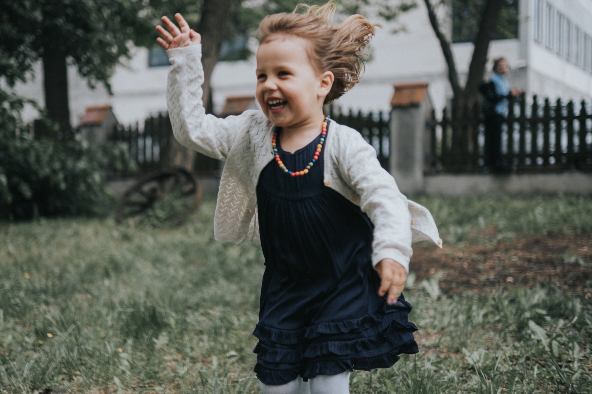 Rozesmiana dziewczynka w granatowej sukience z falbankami i kolorowym naszyjnikiem biegnie po trawie, machając ręką, z rozwianymi włosami.