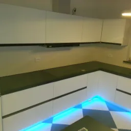 Nowoczesna kuchnia z białymi frontami szafek, czarnym blatem i podświetleniem LED w kolorze niebieskim u dołu szafek, czarno-biała podłoga.