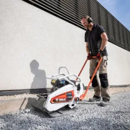 Pracownik w słuchawkach obsługuje zagęszczarkę płytową Bobcat F15.50 na żwirowym podłożu, widoczny cień urządzenia na jasnej ścianie z poziomymi żaluzjami w tle.