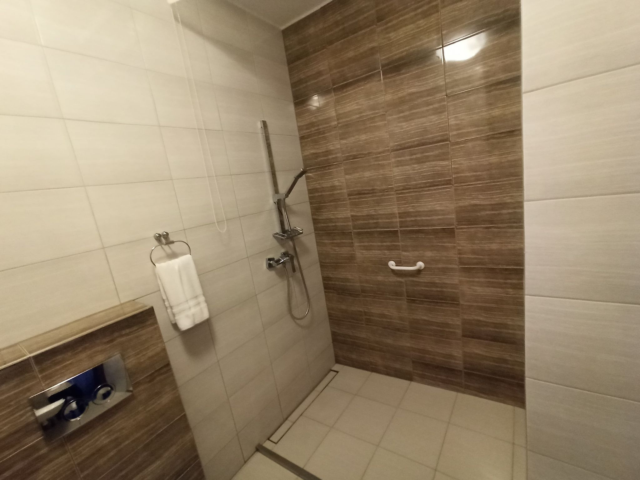 Nowoczesna kabina prysznicowa z brązowo-beżowymi płytkami imitującymi drewno, chromowana armatura i uchwyt dla niepełnosprawnych.