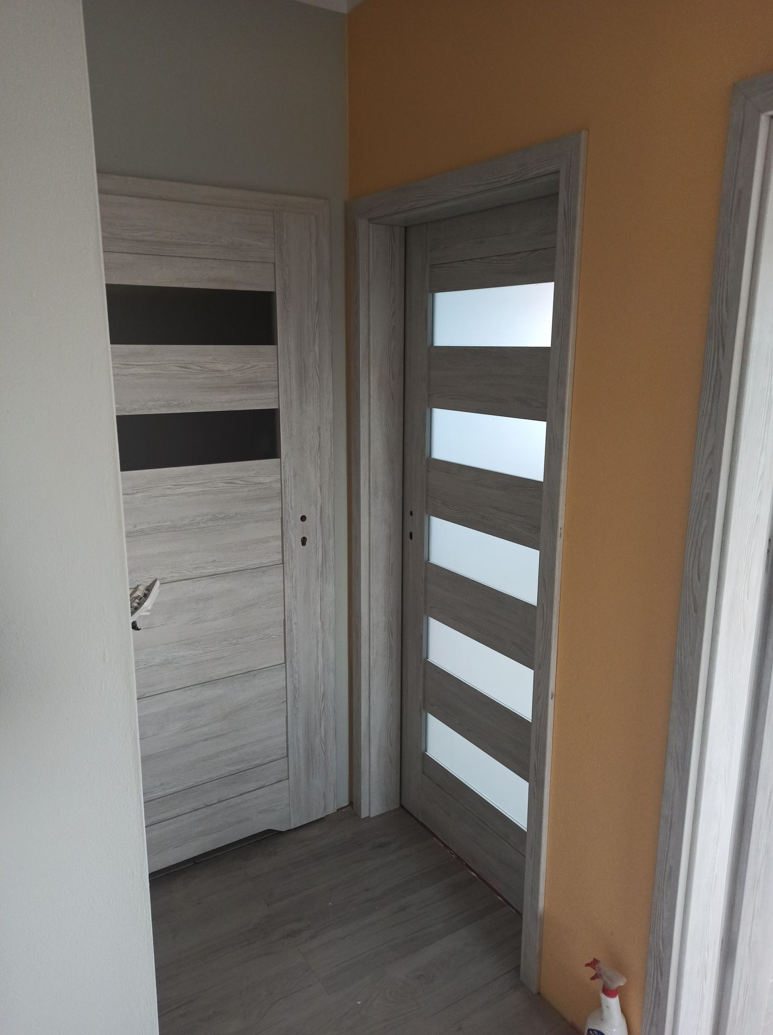 Dwa rodzaje szarych drzwi wewnętrznych z przeszkleniami, jeden z poziomymi pasami matowego szkła, drugi z pionowymi panelami, zamontowane w odnowionym mieszkaniu z szarą podłogą i pomarańczową ścianą.