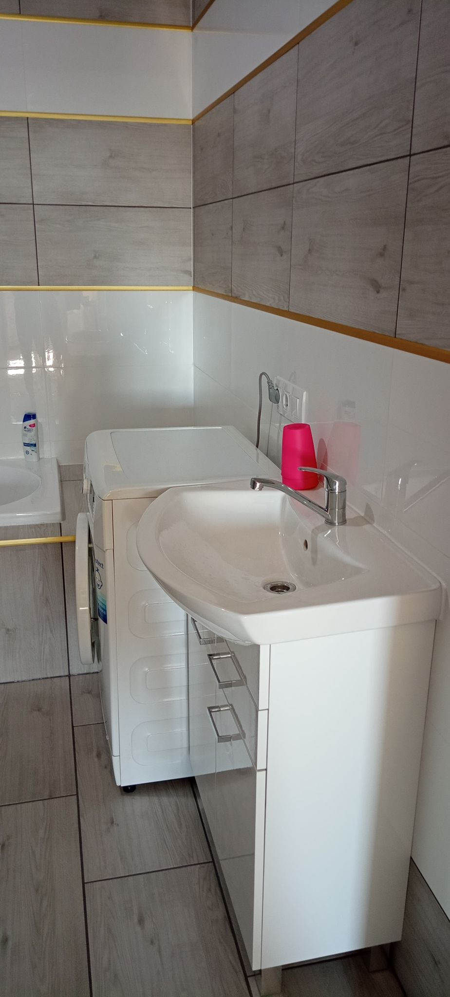 Umywalka nablatowa z szafką w małej łazience, obok pralka, szaro-białe płytki imitujące drewno z złotymi listwami.