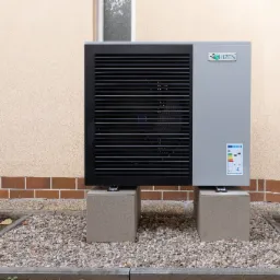 Zewnętrzna jednostka pompy ciepła Shizen na betonowych podstawach, z widoczną etykietą energetyczną A+++, umieszczona przy elewacji budynku z ceglanym cokołem i kamienistym podłożem.