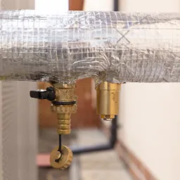 Izolowany przewód z zaworem zapobiegającym zamarzaniu i drugim elementem, prawdopodobnie odpowietrznikiem, w instalacji grzewczej. Widoczny czarny uchwyt zaworu i mosiężna zatyczka na sznurku.