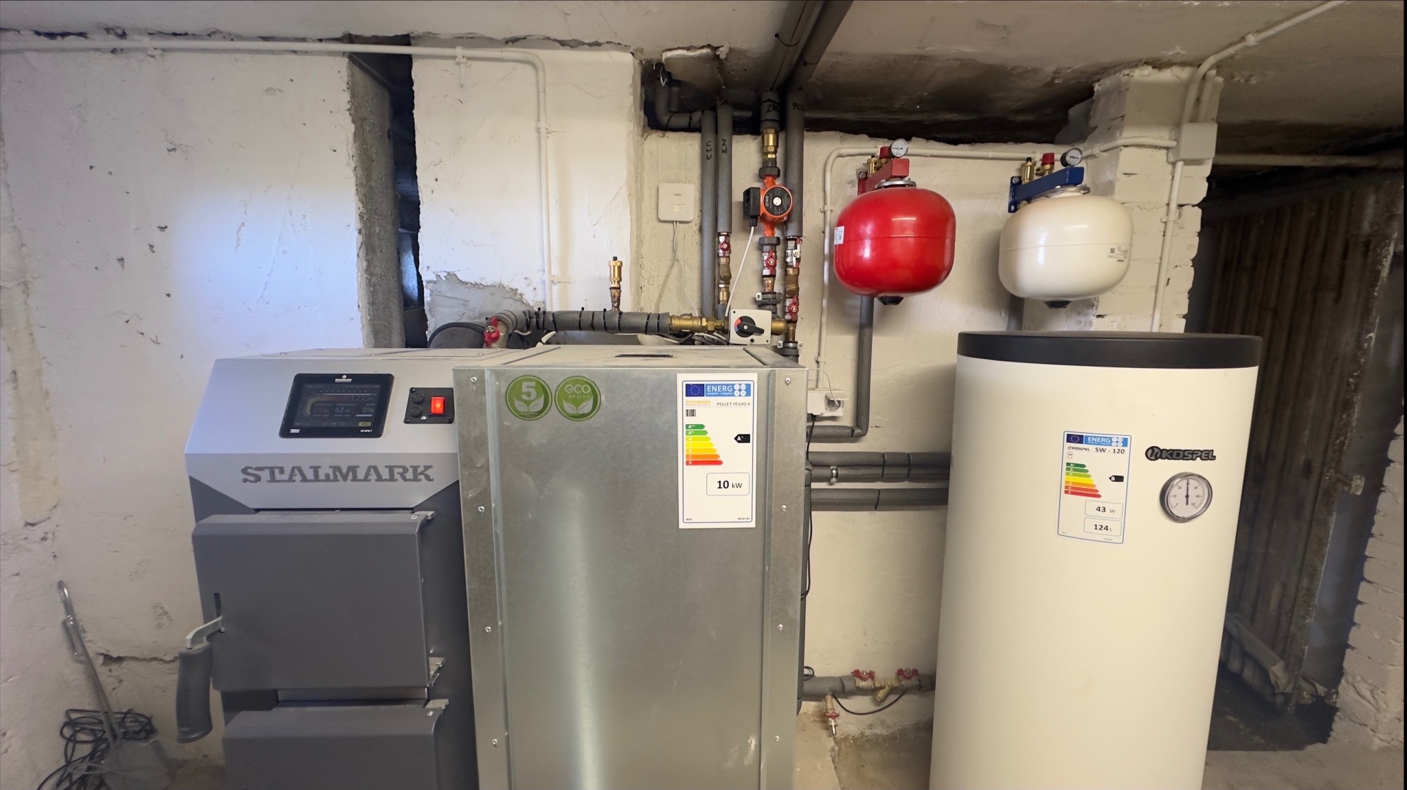 Zestaw kotła na paliwo stałe marki Stalmark z zasobnikiem Kospel SW-120 w piwnicy, widoczne naklejki informacyjne o klasie energetycznej i parametrach technicznych.