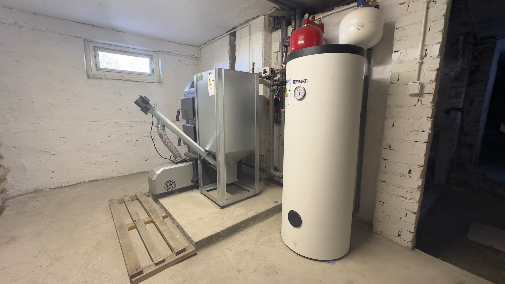 Kotłownia w piwnicy z piecem na paliwo stałe z podajnikiem i bojlerem Kospel, widoczna paleta na podłodze.