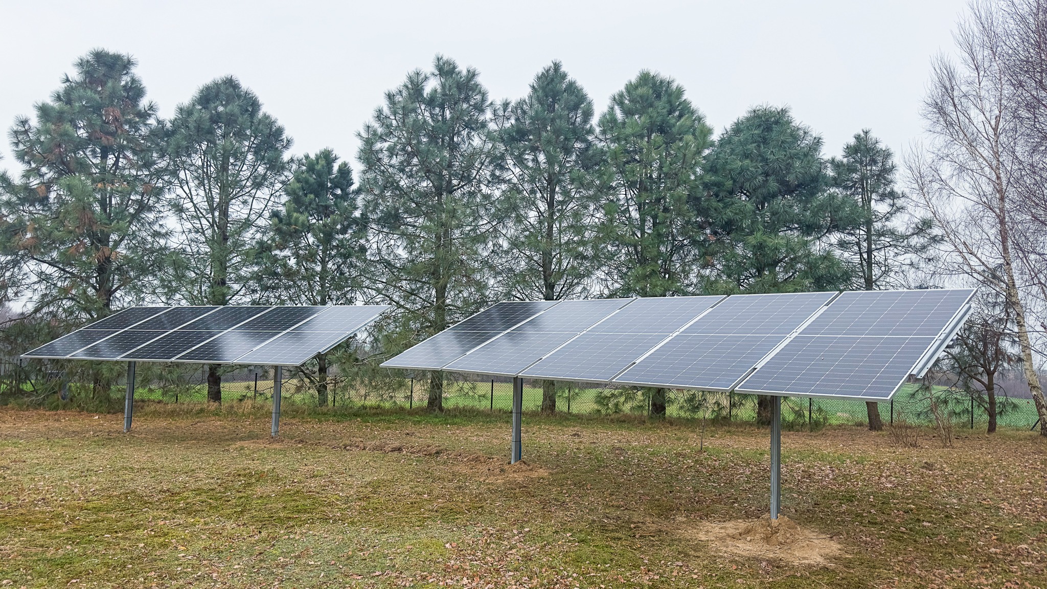 Dwa panele słoneczne na metalowych stelażach, zainstalowane na trawiastym terenie z drzewami iglastymi w tle, widoczne ślady po montażu wokół podstawy stelaży.