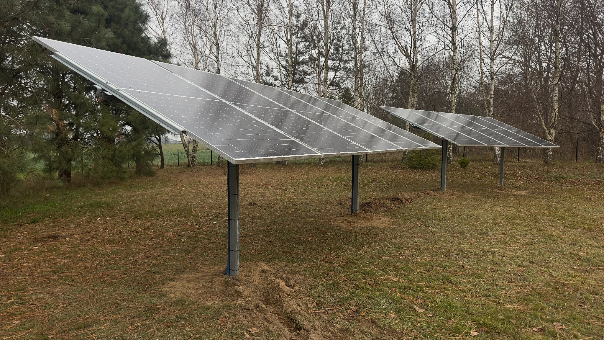 Dwa panele słoneczne zamontowane na metalowych słupach na zewnątrz, z widocznymi śladami po pracach ziemnych i drzewami w tle.