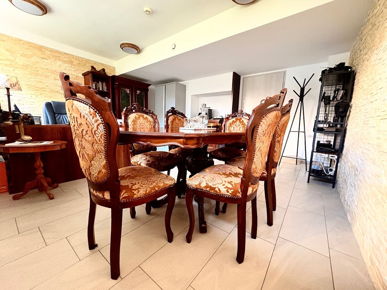 Elegancka jadalnia z ciemnymi, zdobionymi krzesłami i stołem. W tle biurko z lampą i regał. Na ścianie dekoracyjna okładzina z kamienia.