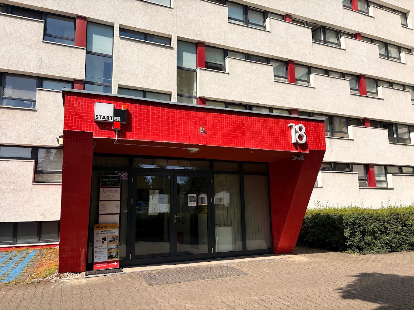 Wejście do budynku z czerwonymi elementami i numerem 18. Na drzwiach tabliczki informacyjne. Widoczny szyld STARTER i fragment elewacji z oknami.