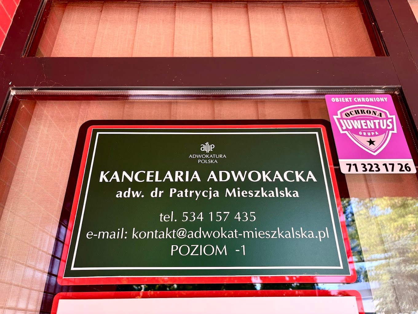 Tabliczka informacyjna Kancelarii Adwokackiej adw. dr Patrycji Mieszkalskiej na drzwiach, obok naklejka firmy ochroniarskiej Juwentus. Widok z zewnątrz.