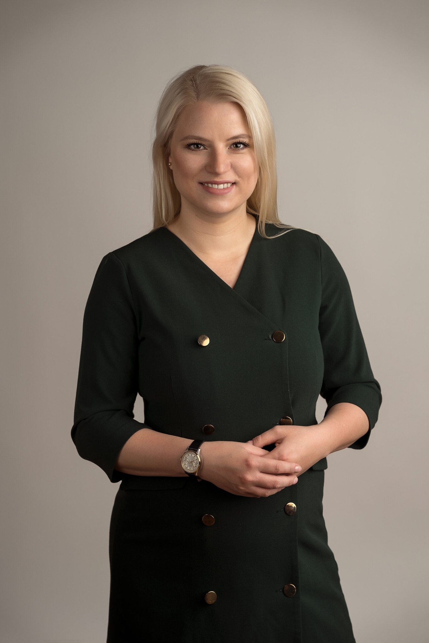 Portret uśmiechniętej blondynki w ciemnozielonej sukience z guzikami i zegarkiem, na neutralnym tle. Profesjonalny wizerunek prawniczki.