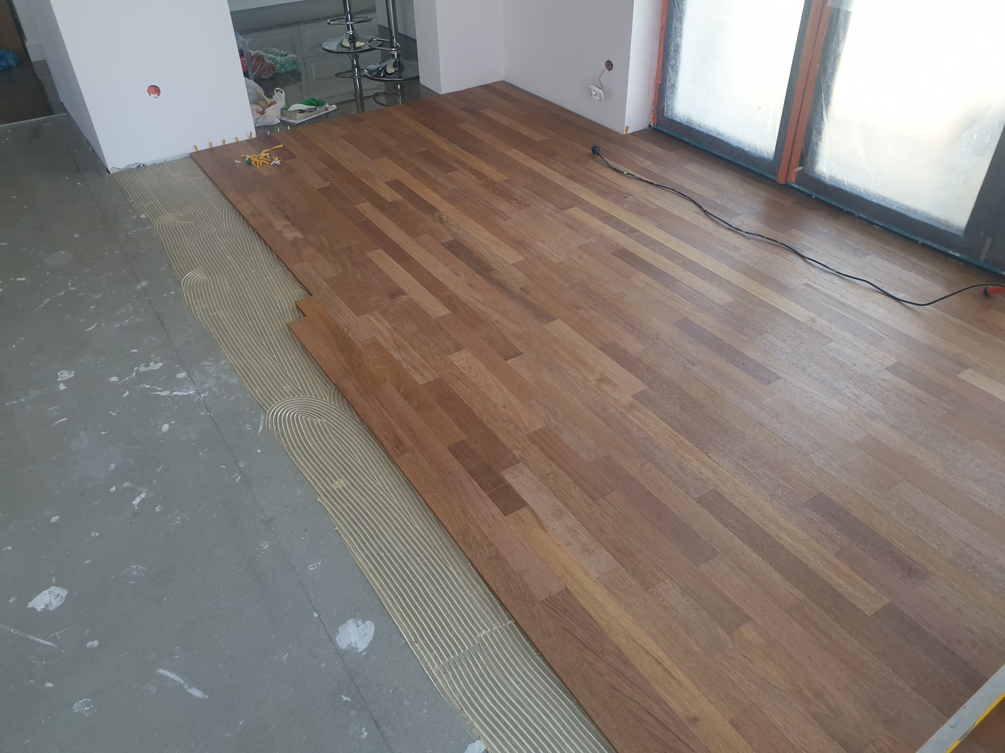 Proces układania parkietu: widoczny fragment podłogi z nałożonym klejem w charakterystyczne paski, sąsiadujący z ułożonymi, lakierowanymi deskami parkietowymi o ciepłym, brązowym odcieniu, w tle...