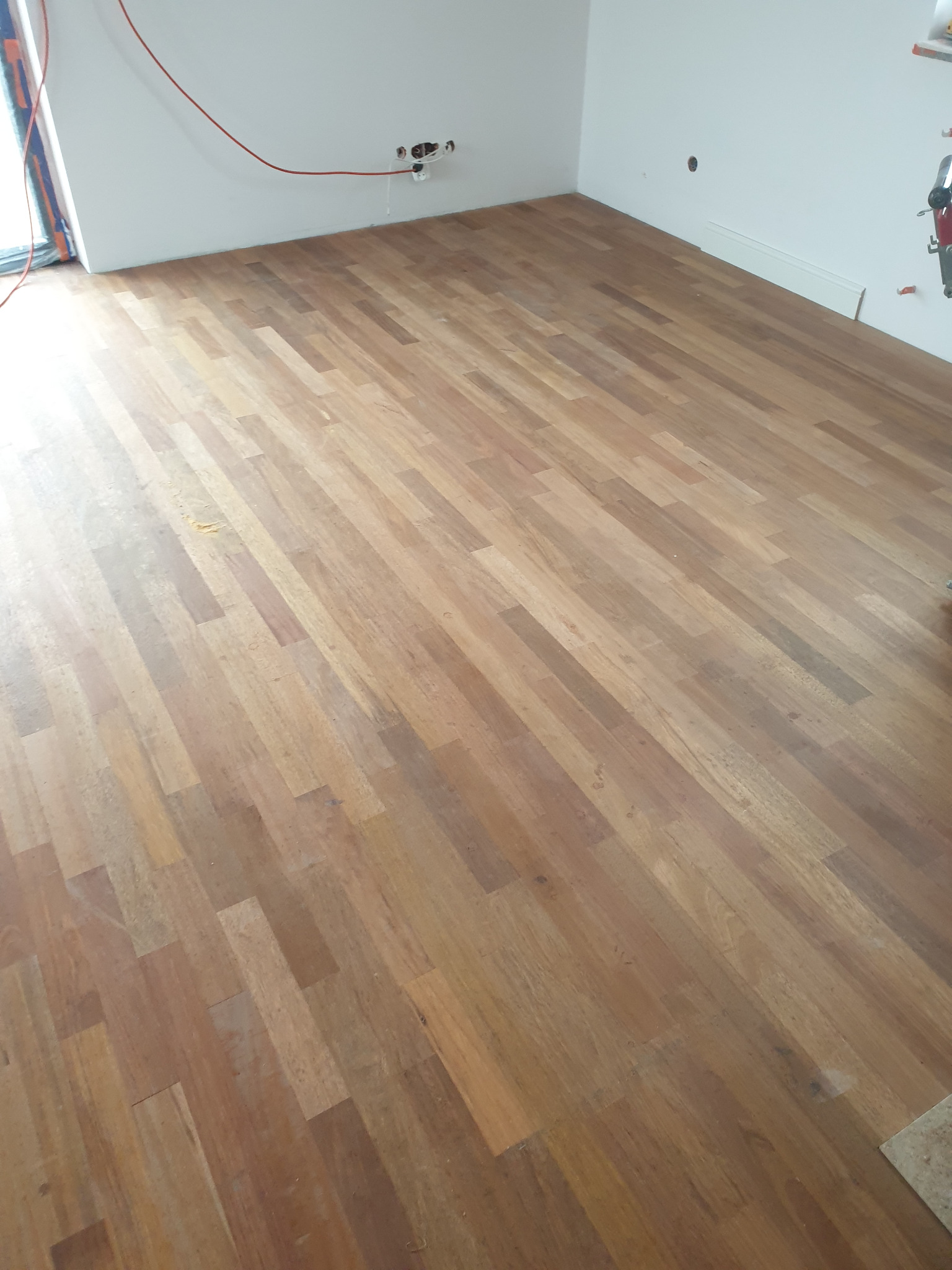 Świeżo położony parkiet z drewna egzotycznego w jasnym, pustym pomieszczeniu z widocznymi instalacjami elektrycznymi na ścianie.