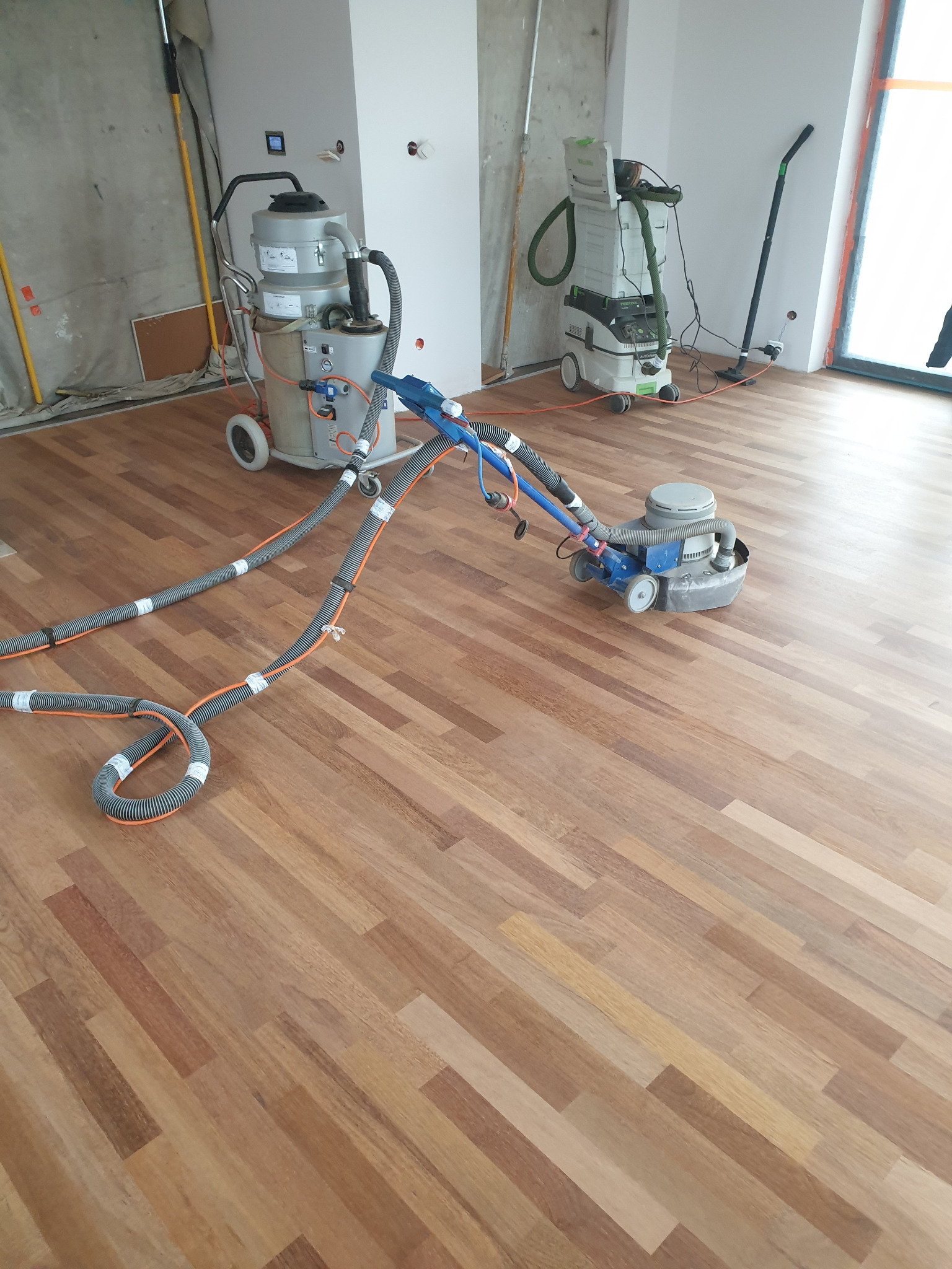 Parkiet z drewna egzotycznego szlifowany maszyną z podłączonym odkurzaczem przemysłowym, widoczne przewody i drugi odkurzacz w tle, surowe ściany w trakcie remontu.