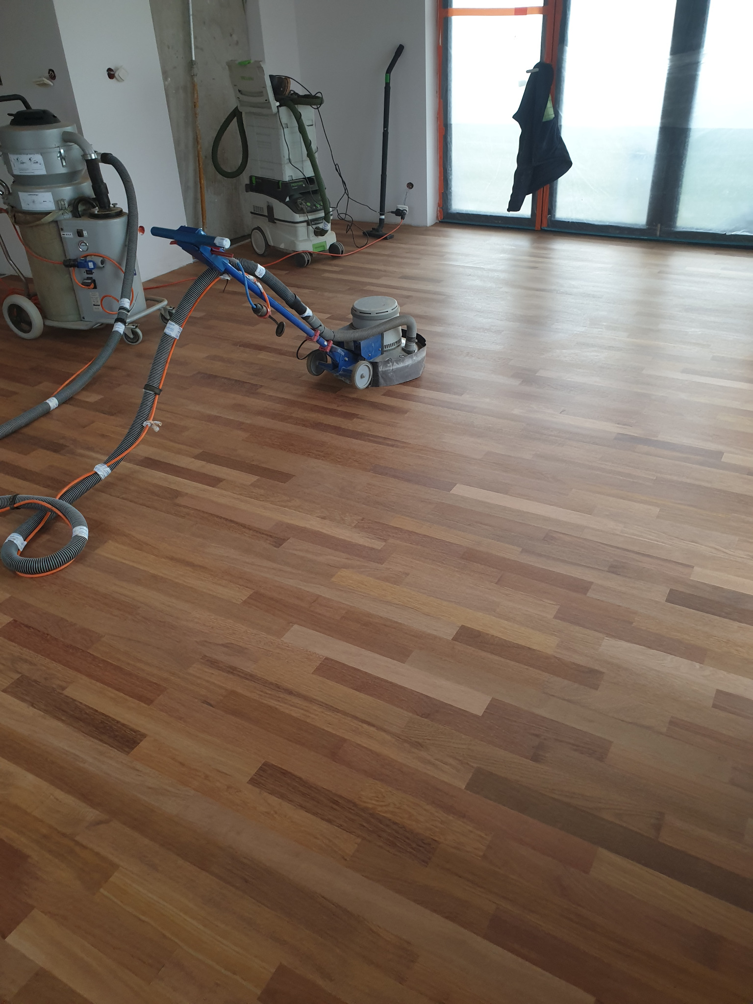 Cyklinowanie parkietu w trakcie realizacji, widoczne maszyny do cyklinowania i odkurzacz przemysłowy, parkiet w odcieniach brązu, duże okno z widokiem na zewnątrz.