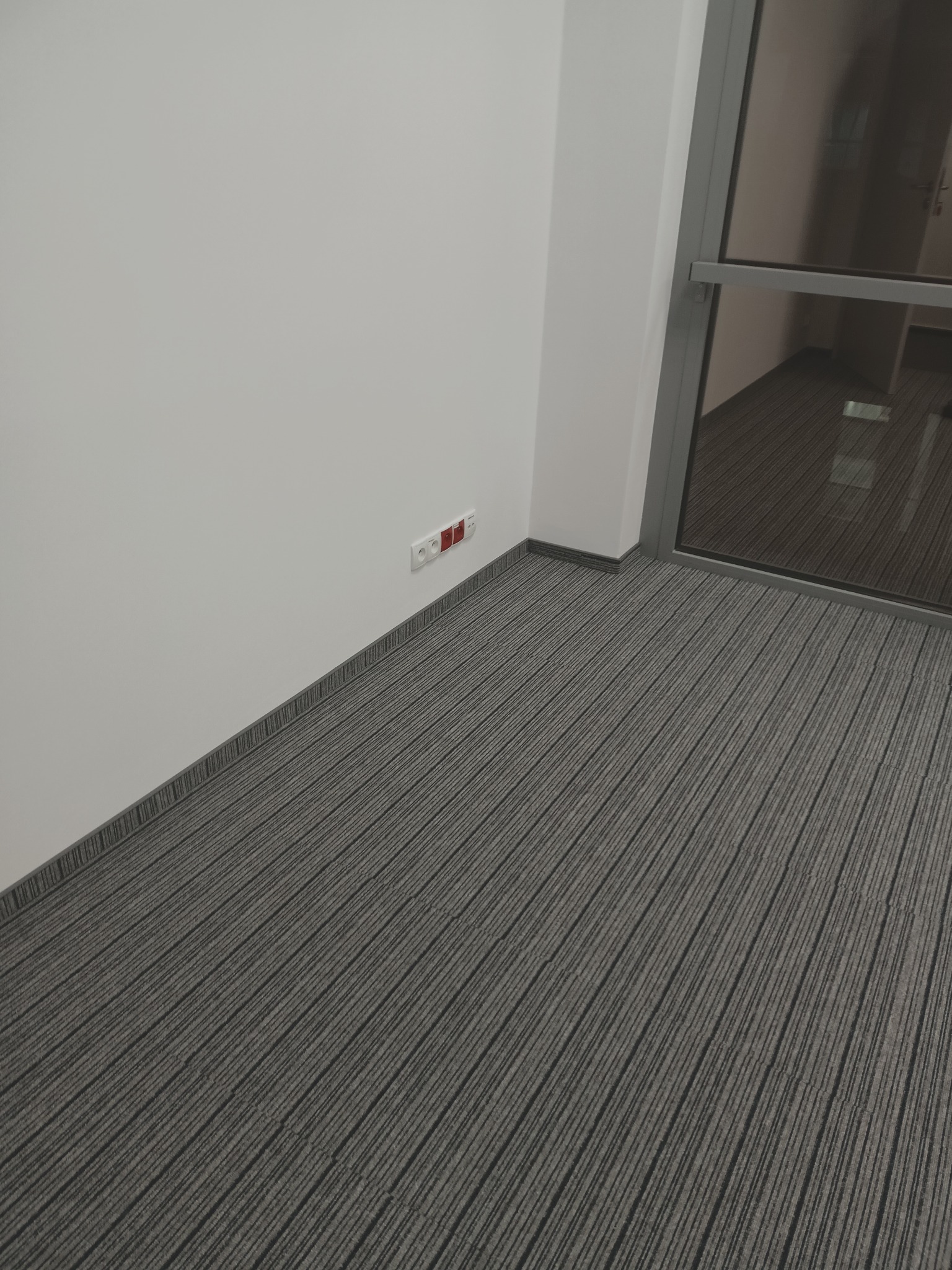 Szara wykładzina dywanowa w biurze z widocznymi gniazdkami elektrycznymi na ścianie i szklanymi drzwiami.