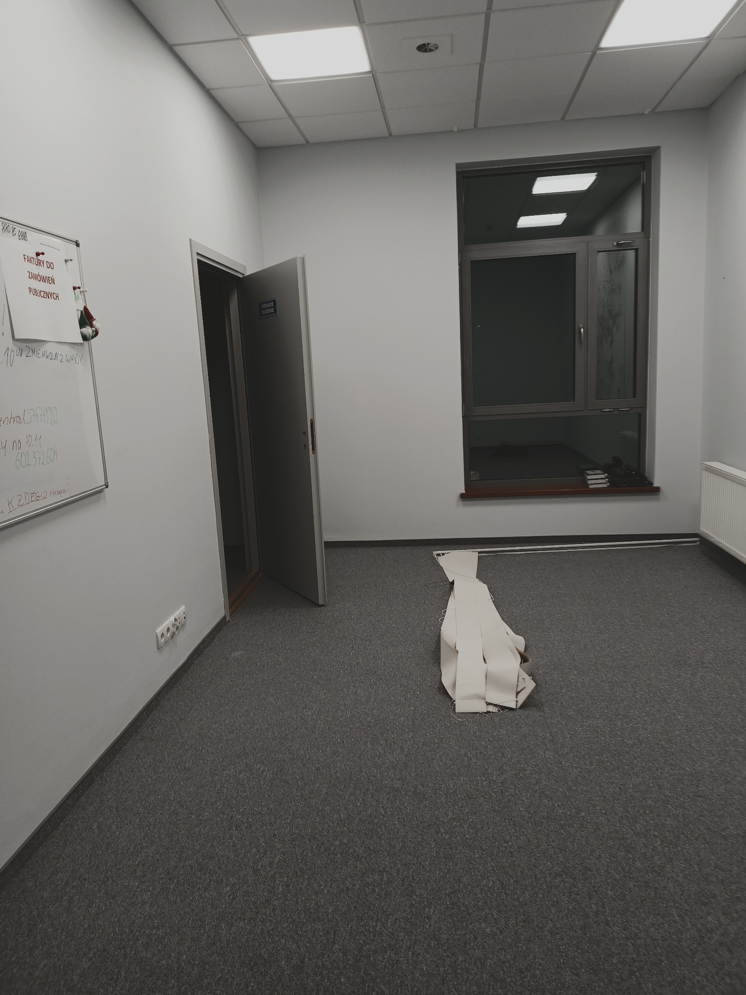 Przygotowanie do ułożenia wykładziny w biurze: rozłożone pasy materiału na szarej podłodze, widoczna tablica suchościeralna z notatkami i częściowo otwarte drzwi.