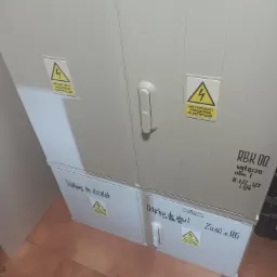 Dwu-segmentowa, biała szafa elektryczna z naklejkami ostrzegawczymi o napięciu, umieszczona w pomieszczeniu z brązową podłogą.