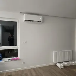 Biały klimatyzator ścienny z wyświetlaczem ustawionej temperatury, zamontowany w nowoczesnym wnętrzu z drewnianą podłogą, białą ścianą, grzejnikiem i oknem z widokiem na miasto nocą.