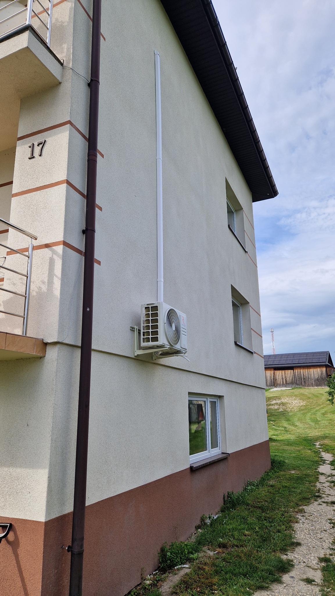 Zewnętrzna jednostka klimatyzacji zamontowana na elewacji dwupiętrowego domu z numerem 17, widoczna rura odprowadzająca skropliny, w tle budynek gospodarczy z panelami fotowoltaicznymi.
