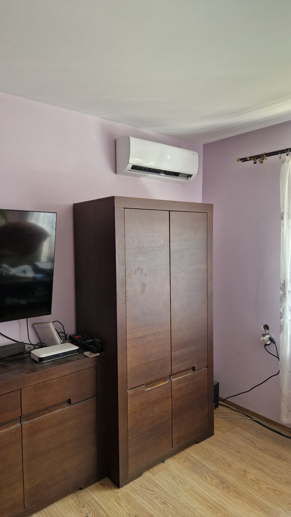 Biała jednostka wewnętrzna klimatyzatora zamontowana nad drewnianą szafą w pokoju o fioletowych ścianach.
