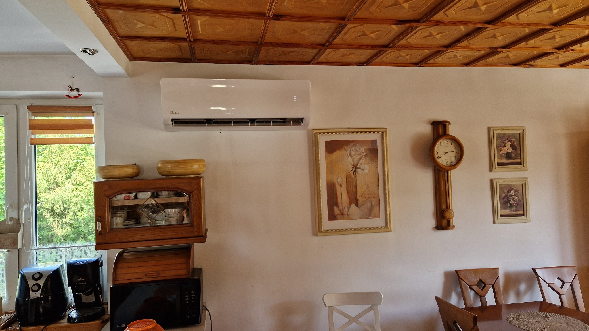 Biała jednostka wewnętrzna klimatyzacji Midea zamontowana na ścianie w pokoju z drewnianym sufitem kasetonowym, obok wiszą obrazy i drewniany zegar.