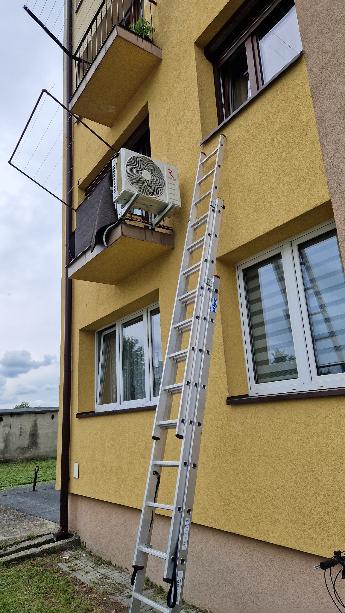 Zewnętrzna jednostka klimatyzatora zamontowana na balkonie budynku z żółtą elewacją, z przystawioną aluminiową drabiną Krause.