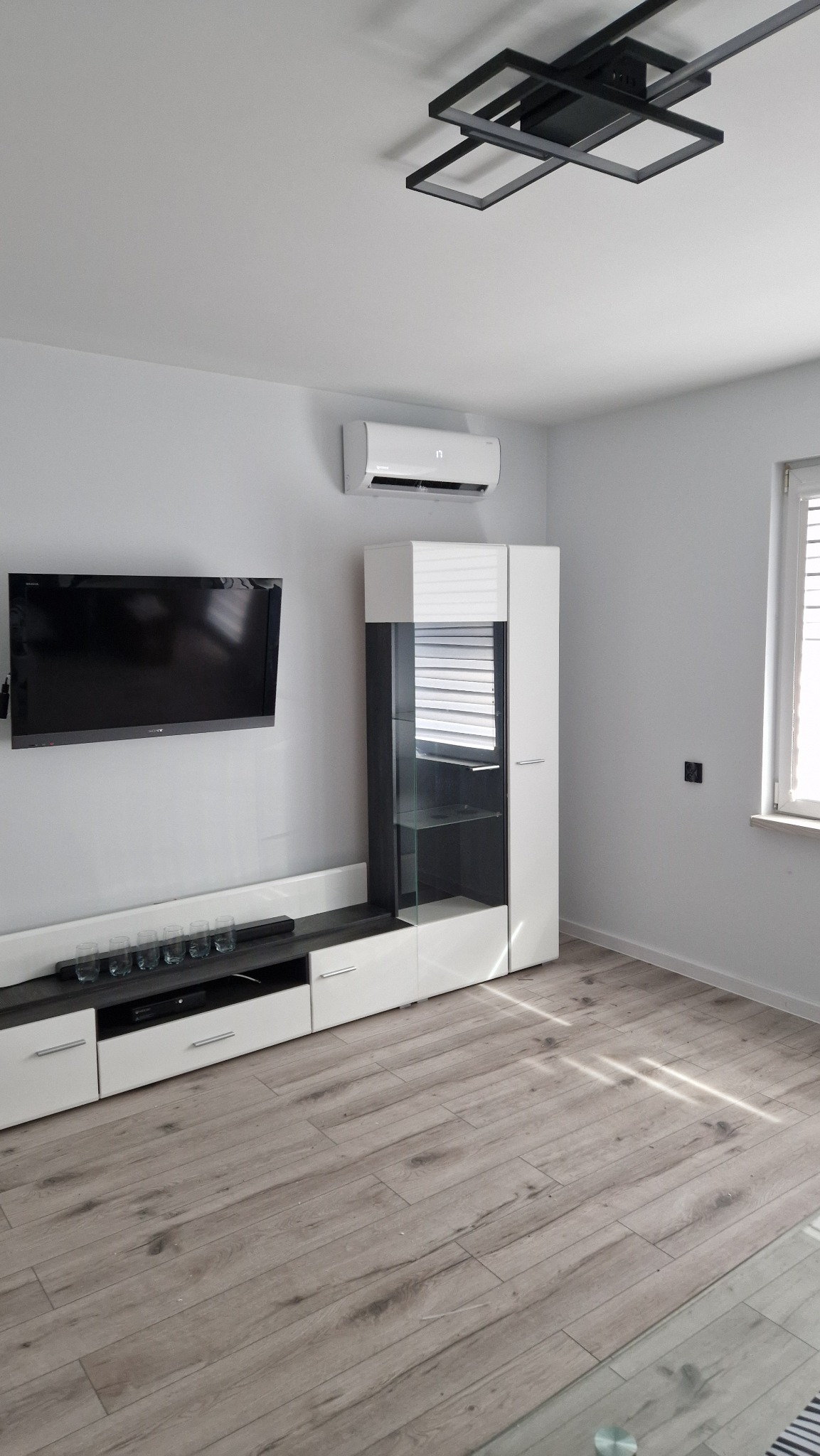 Nowoczesny salon z białym klimatyzatorem ściennym, czarnym telewizorem, biało-szarą szafką RTV, szklaną witryną i geometryczną lampą sufitową.
