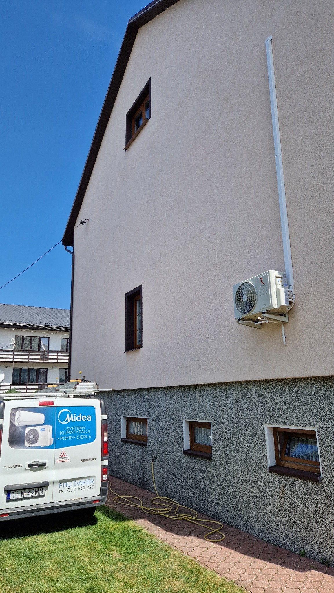 Zewnętrzna jednostka klimatyzacji zamontowana na elewacji domu, widoczny samochód dostawczy z reklamą systemów klimatyzacji i pomp ciepła oraz zwinięty żółty wąż ogrodowy leżący na chodniku z kostki.