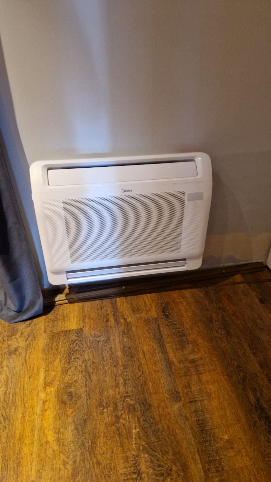 Biała, podłogowa jednostka wewnętrzna klimatyzacji Midea zamontowana przy ścianie, widoczna z perspektywy podłogi z drewnopodobnych paneli.