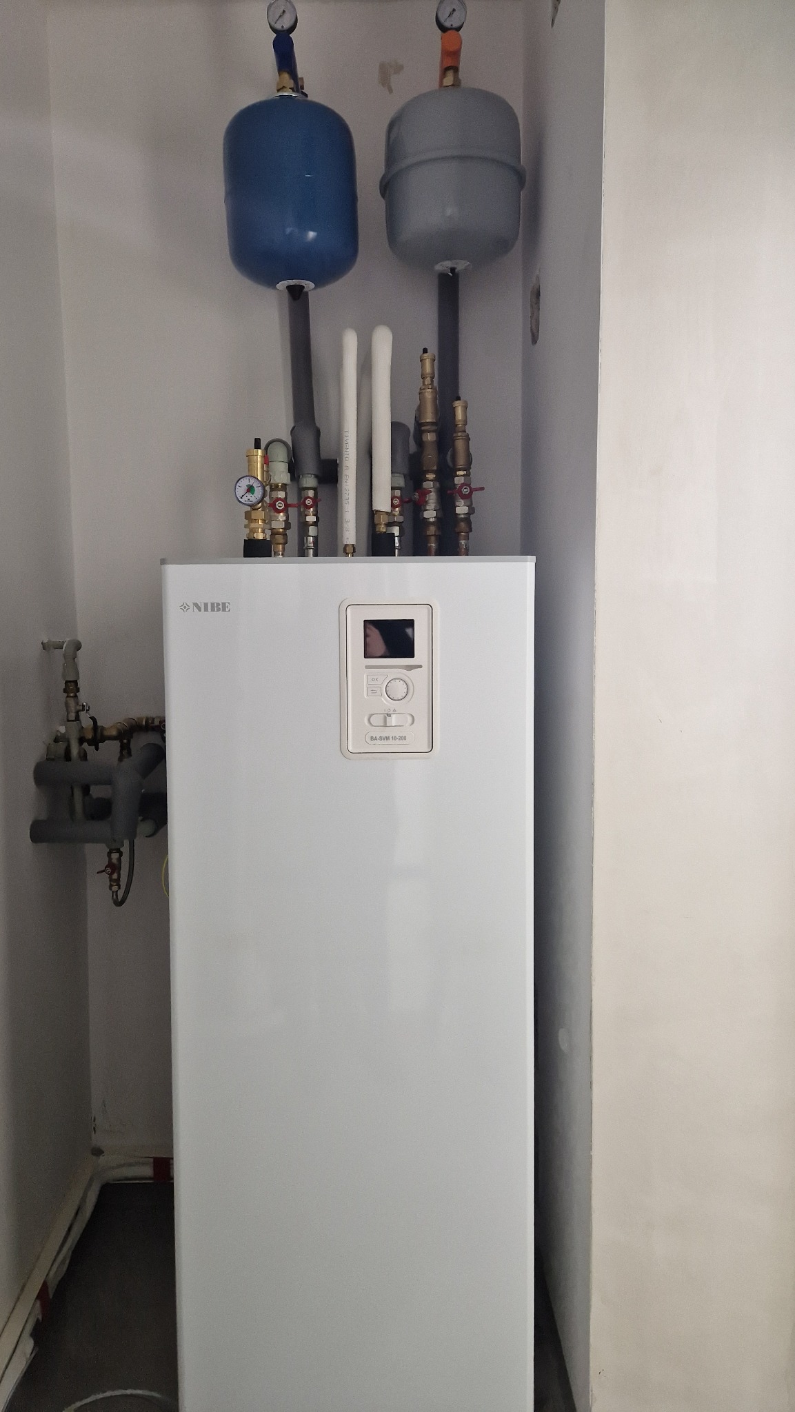 Biała pompa ciepła Nibe z niebieskim i szarym zbiornikiem wyrównawczym, widoczne rury i zawory, zamontowana w rogu pomieszczenia o jasnych ścianach.