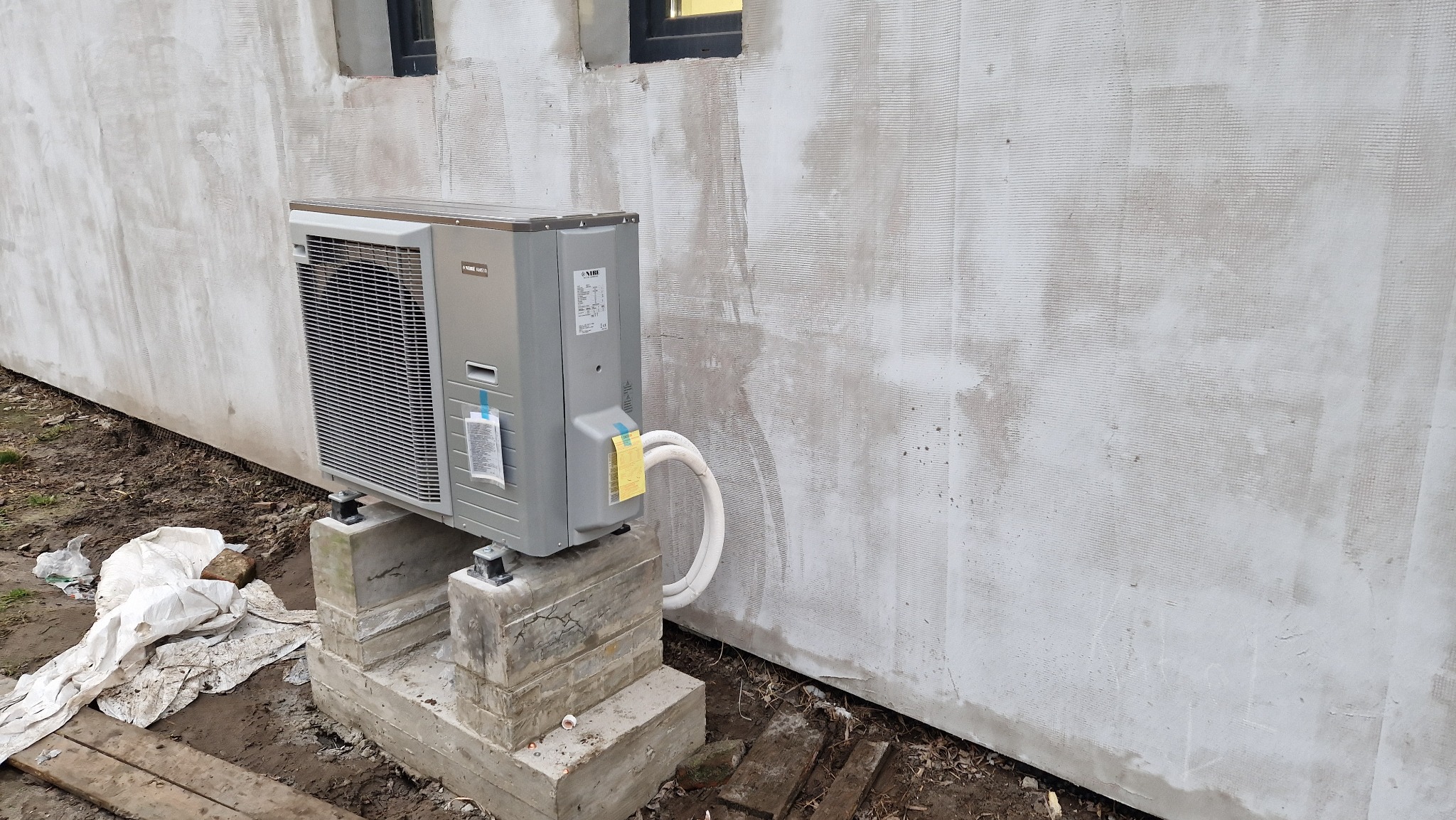 Szara jednostka zewnętrzna klimatyzatora NIBE zamontowana na betonowych bloczkach przy szarej ścianie budynku z widocznymi przewodami chłodniczymi.