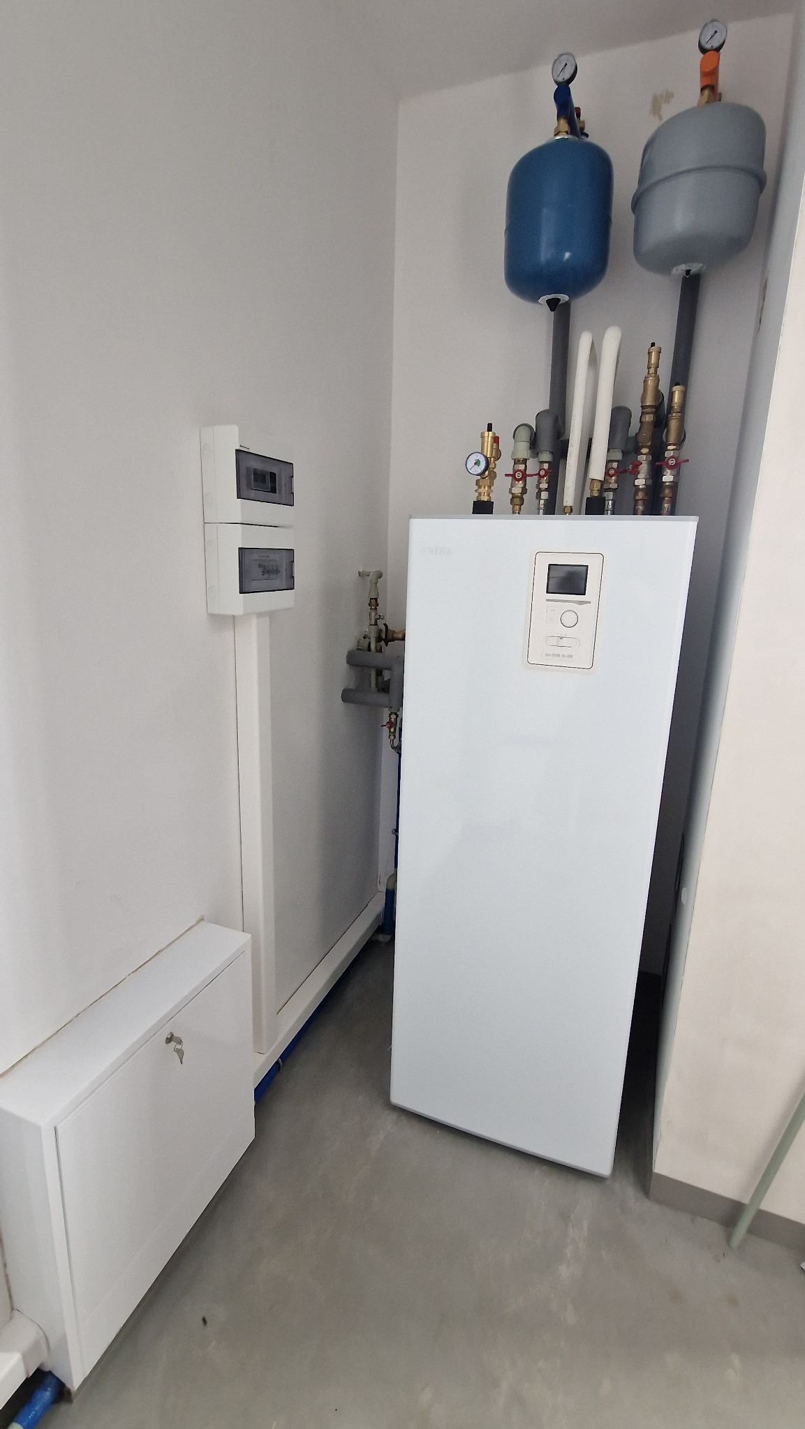 Biała jednostka wewnętrzna z wyświetlaczem, rurami i zbiornikami wyrównawczymi w pomieszczeniu technicznym z szarą posadzką.