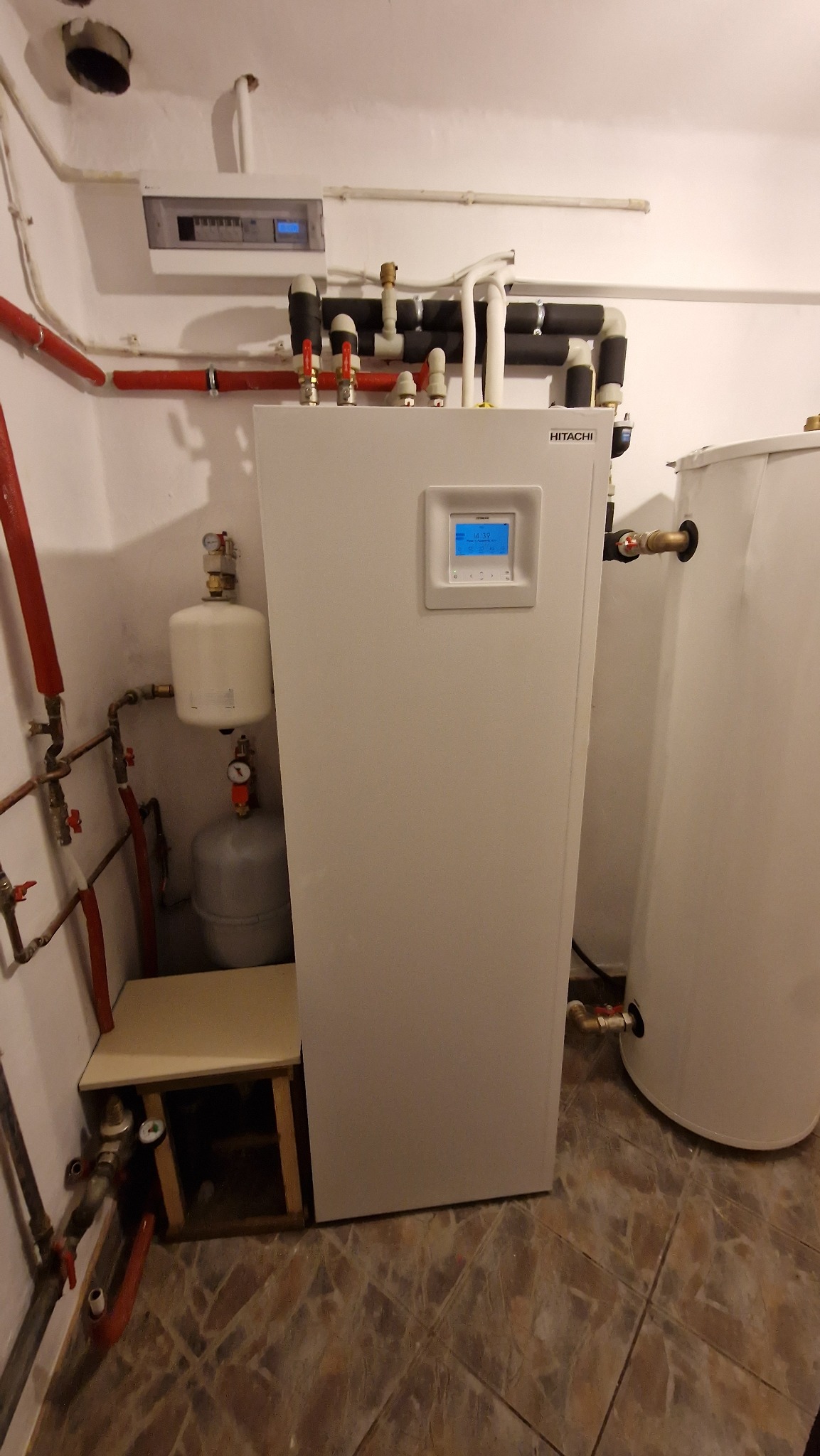 Biała pompa ciepła marki Hitachi z widocznym panelem sterowania i podłączeniami hydraulicznymi w pomieszczeniu technicznym, obok zasobnik ciepłej wody.